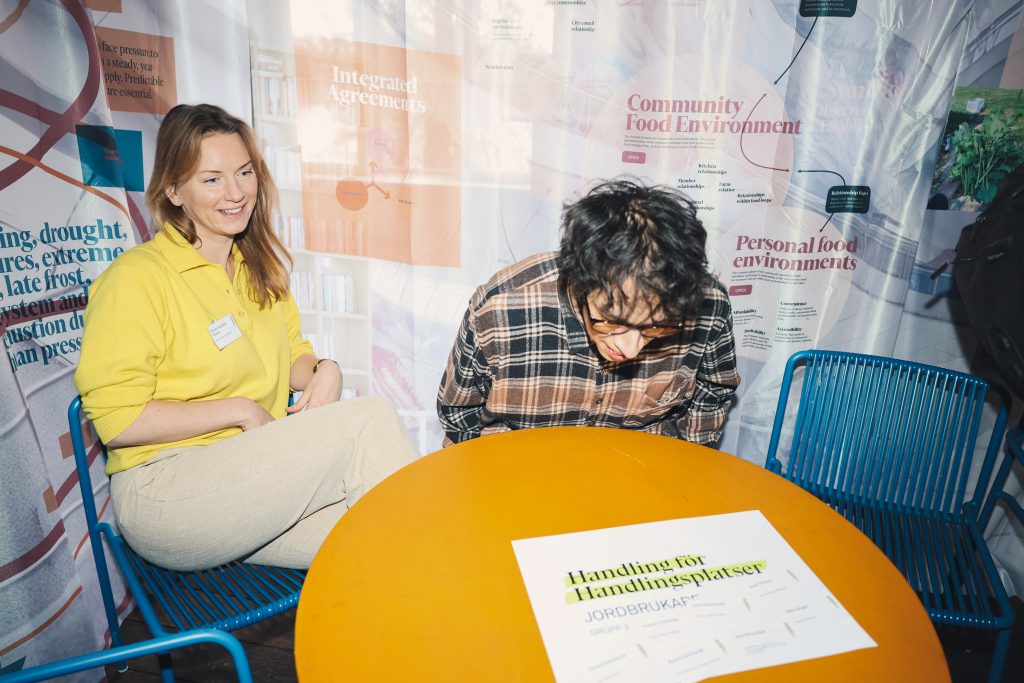 Two people sit at a bright orange round table, woman in a yellow sweater smiling at the camera with a man in a plaid shirt beside her in a booth with colorful wall graphics.