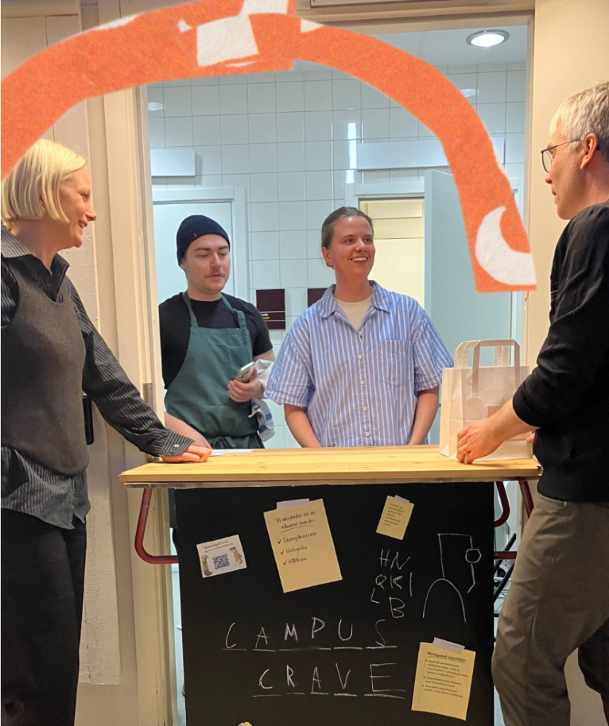 Four people stand around a wooden counter in a campus cafe, smiling, with a chalkboard that reads 'CAMPUS CRAVE' and sticky notes visible below the counter sketch.